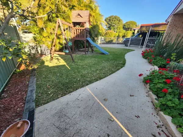 Freshly mown backyard lawn with a curved concrete path, timber cubby house and red flowering garden bed